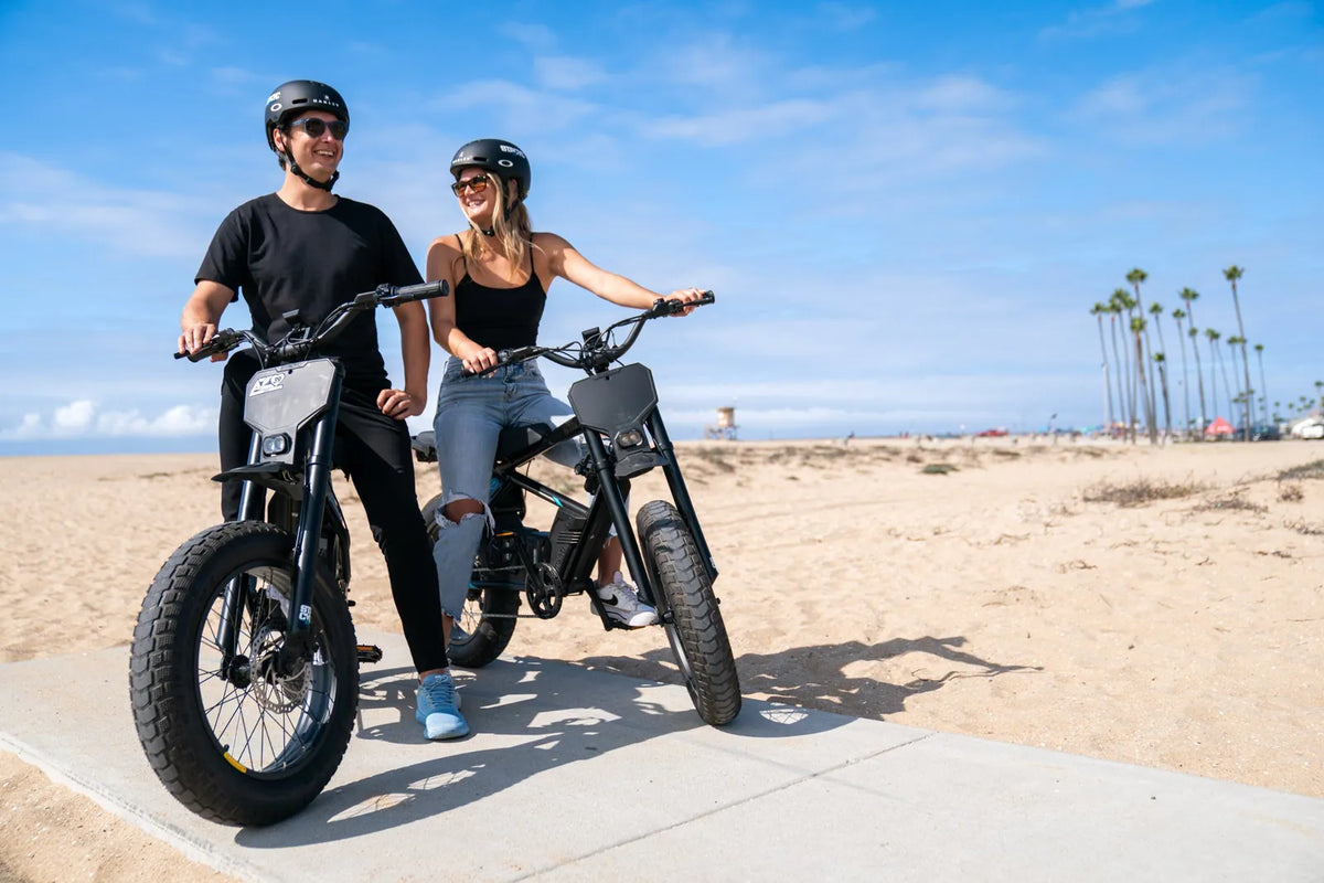 Two riders smiling on STACYC 20hDRIVE e-bikes parked on a beach path