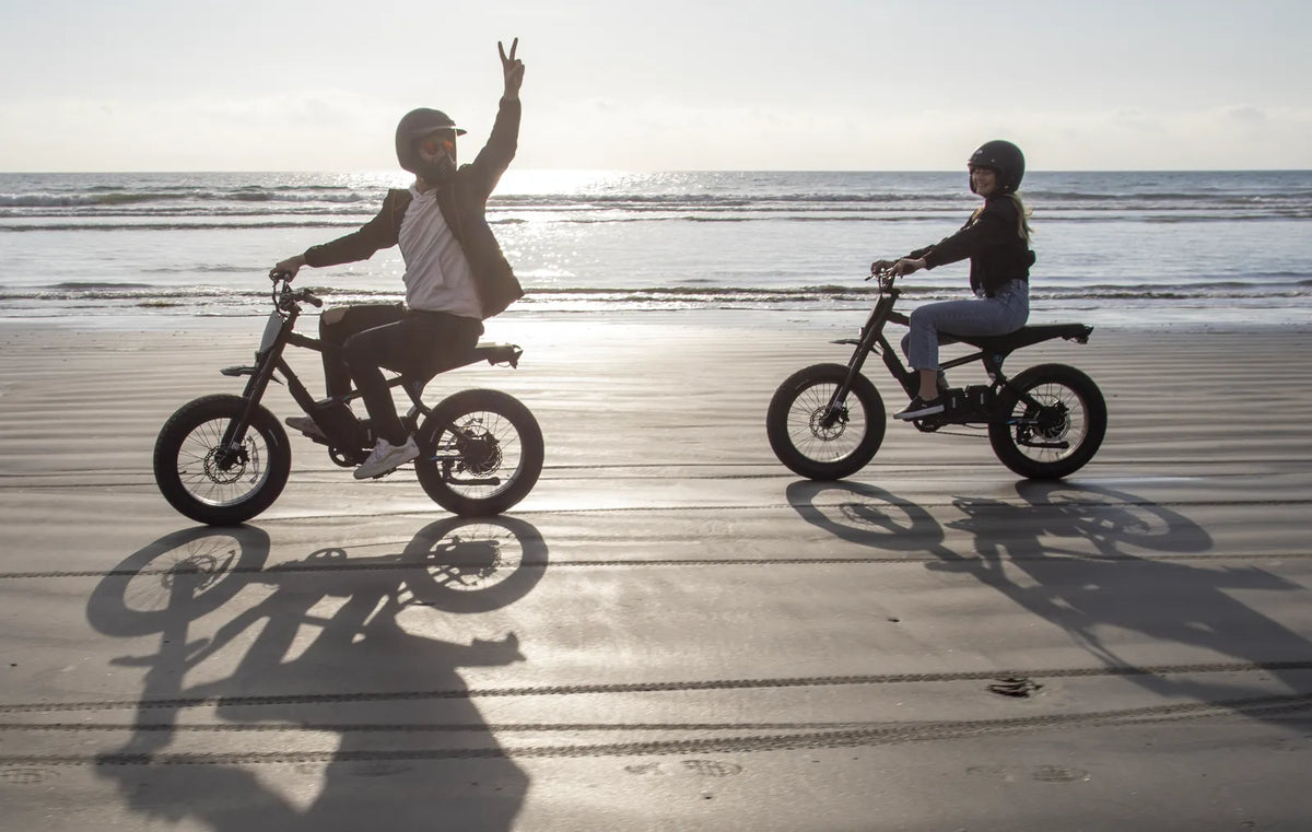 Two riders on STACYC 20hDRIVE e-bikes crossing wet sand at the water's edge, one throwing a peace sign
